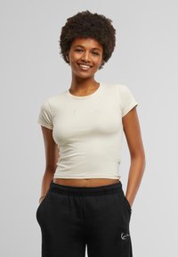 T-shirt cropped beige clair en tissu lisse. Présente des manches courtes et un logo brodé subtil sur la poitrine. Assorti à un pantalon de survêtement noir.