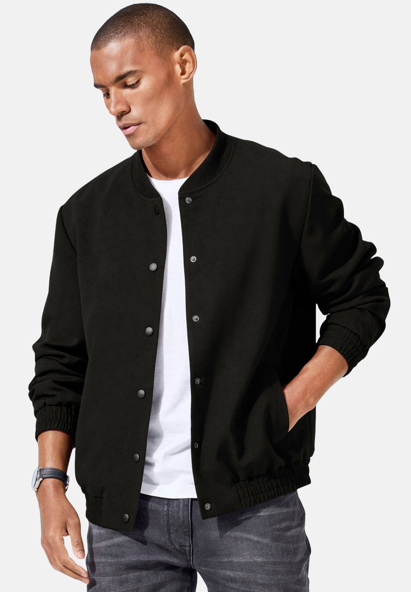 Black bomber jacket with snap buttons, ribbed cuffs, and waistband. Worn over a white shirt and styled with gray jeans. Smooth fabric texture.