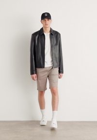 Sort læderjakke, hvid T-shirt, beige shorts, hvide sneakers og en sort cap. Modellen står foran en neutral baggrund.