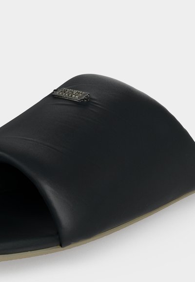 Μαύρη σαγιονάρα slide με φαρδιά επενδυμένη λωρίδα και μικρό ορθογώνιο μεταλλικό διακοσμητικό έμβλημα στην κορυφή, μπεζ σόλα ορατή από κάτω.