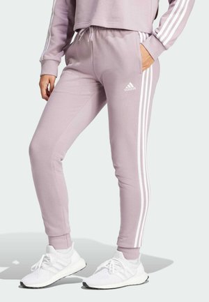 Osoba nosząca jasnofioletowe spodnie dresowe Adidas z białymi paskami i białe sportowe buty, stoi z jedną ręką w kieszeni.