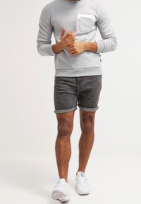 Sudadera gris con un bolsillo en el pecho, combinada con pantalones cortos de denim negros enrollados. Zapatillas blancas completan el conjunto.