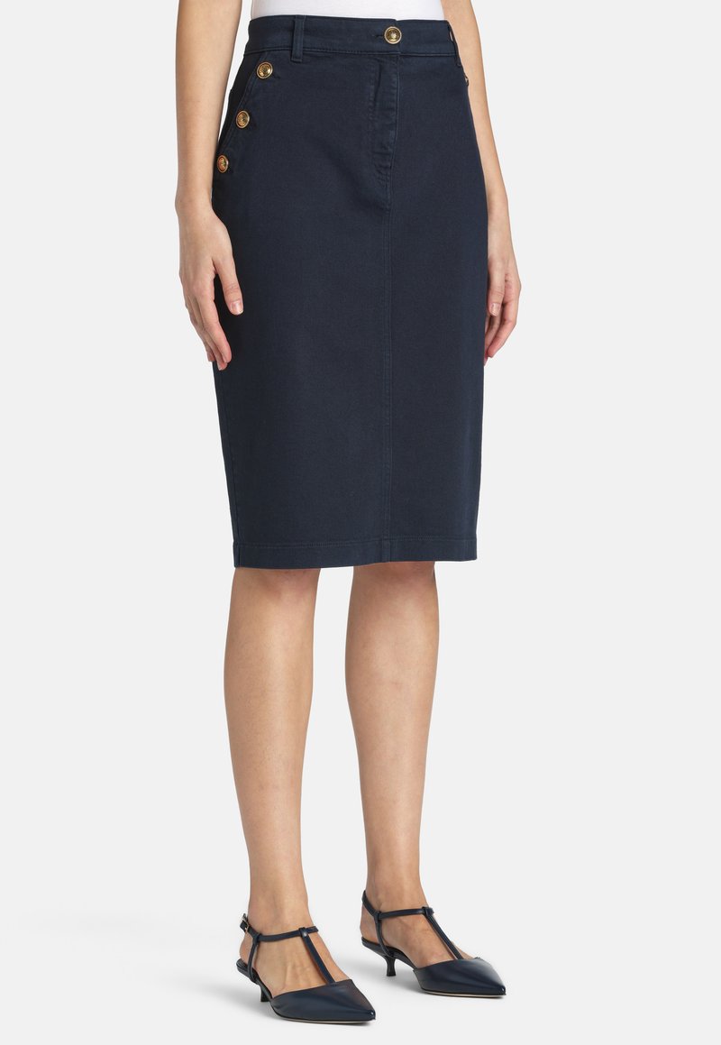 Femme portant une jupe crayon bleu marine au genou avec des boutons dorés sur la ceinture et sur le côté, associée à des escarpins à bride en T bleu marine.