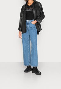 Schwarzes Kunstlederhemd über einem schwarzen T-Shirt, kombiniert mit hellblauen weiten Jeans und schwarzen klobigen Stiefeletten.