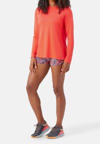 Dlouhý rukáv, jasně oranžový sportovní top spárovaný s kraťasy s květinovým vzorem. Černé běžecké boty s růžovými a oranžovými akcenty. Hladké, lehké materiály.