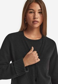 Czarny kardigan z dekoltem w szpic, ściągaczami przy mankietach oraz zapięciem na guziki, noszony na dopasowanej czarnej bluzce. Miękka, teksturowana dzianina.