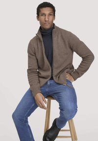 Brauner Strickjacke mit Reißverschluss und gerippten Bündchen, getragen über einem marineblauen Rollkragenpullover, kombiniert mit blauen Jeans und schwarzen Schuhen, sitzt auf einem Holzstuhl.