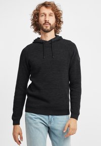 Schwarze, strukturierte Kapuzenjacke mit Kordelzug, gerippten Bündchen und Saum. Getragen über hellblauen Jeans. Runder Logo-Patch auf der linken Schulter.