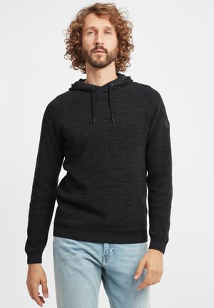 Schwarze, strukturierte Kapuzenjacke mit Kordelzug, gerippten Bündchen und Saum. Getragen über hellblauen Jeans. Runder Logo-Patch auf der linken Schulter.