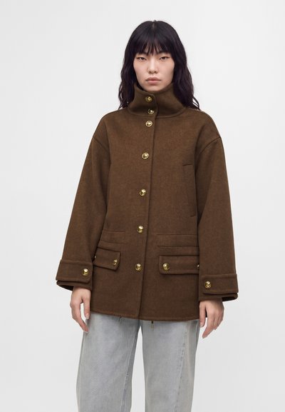 Femme portant un manteau marron à col haut avec boutons dorés et poches avant, associé à un pantalon gris clair, debout devant un fond uni.