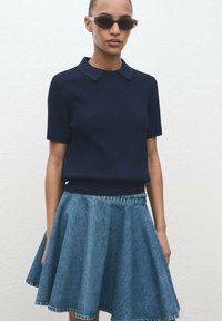Haut en tricot à manches courtes de couleur marine avec un col, associé à une jupe en denim A-line bleu clair. Le mannequin porte des lunettes de soleil noires sur un fond neutre.