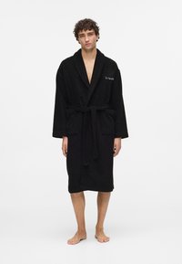 Svart frottébadekåpe med sjalkrage, omslag foran og belte i livet. Har en lommepatch og broderte detaljer på brystet.