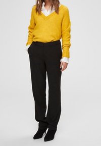 Żółty sweter w serek wykonany z miękkiego materiału, połączony z czarnymi dopasowanymi spodniami i czarnymi szpilkami. Outfit podkreśla kontrast i warstwowanie.