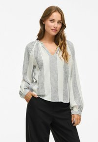 Femme aux cheveux longs portant une blouse blanche à rayures verticales bleues et un pantalon noir, se tenant avec une main dans la poche.