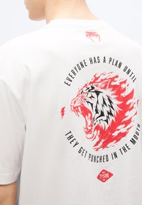 Λευκό βαμβακερό μπλουζάκι με τολμηρό κόκκινο και μαύρο γραφικό λιονταριού με φλόγες. Το κείμενο γράφει, «Όλοι έχουν ένα σχέδιο μέχρι να φάνε μια γροθιά στο στόμα.»