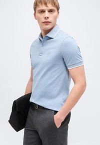 Jeune homme portant un polo bleu clair et un pantalon gris, tenant une veste noire dans une main, regardant légèrement sur le côté.