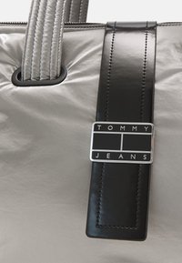 Hopeapintainen laukku, jossa on sileä pinta ja kontrastivärinen musta hihna, ja hopeinen logo-levy, jossa on kohokuviolla teksti "TOMMY JEANS."
