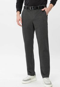 Mann trägt dunkelgraue Hose, schwarzen Gürtel, schwarzes Langarmshirt und weiße Sneakers und steht mit einer Hand in der Tasche.