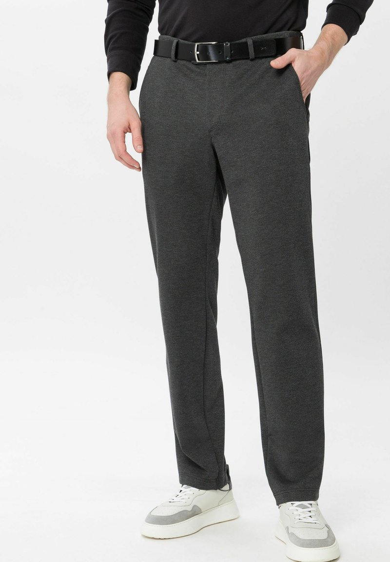 Mann trägt dunkelgraue Hose, schwarzen Gürtel, schwarzes Langarmshirt und weiße Sneakers und steht mit einer Hand in der Tasche.