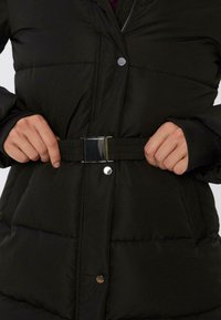 Sort pufferjakke med en blank spænde-bælte, trykknapper og en quiltet tekstur. Ærmerne har en tætsiddende design for ekstra varme.