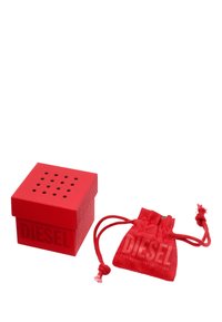 Scatola quadrata rossa con finitura testurizzata e coperchio perforato, con la parola "DIESEL" in rilievo; accompagnata da un sacchetto rosso abbinato con coulisse.