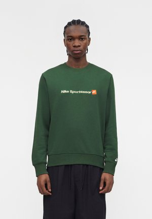 Mladý muž s vrkočmi, oblečený v zelenej mikine Nike Sportswear a čiernych nohaviciach, stojaci pred obyčajným bielym pozadím.