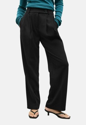 Schwarze plissierte Hosen mit hoher Taille, seitlichen Taschen und einem Knopfverschluss, kombiniert mit schwarzen Absatz-Flip-Flops und einem teal-farbenen Pullover.
