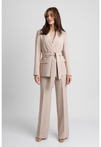 Beżowy dopasowany garnitur z szerokimi spodniami i żakietem z paskiem. Żakiet ma kołnierz szalowy oraz przednie kieszenie naszywane. Gładka tekstura materiału.