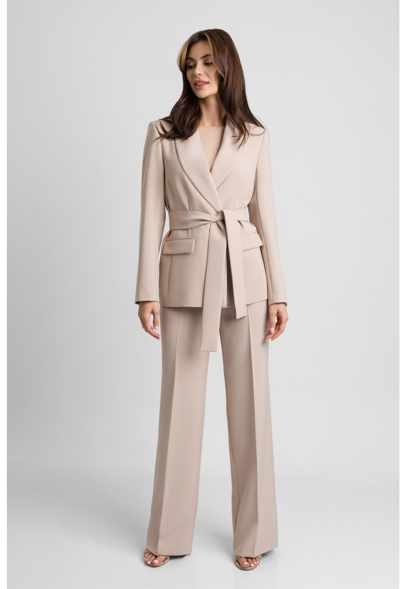 Beżowy dopasowany garnitur z szerokimi spodniami i żakietem z paskiem. Żakiet ma kołnierz szalowy oraz przednie kieszenie naszywane. Gładka tekstura materiału.