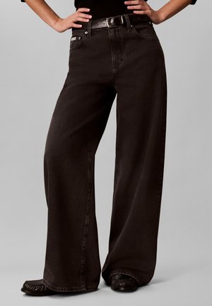 Person iført sorte, højtaljede, vidde jeans med sort bælte og mørke mokkasiner, hænderne hvilende på hofterne mod en grå baggrund.