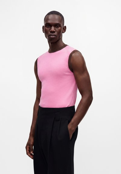 Homme portant un haut rose sans manches avec une bordure marron et un pantalon noir plissé taille haute, debout avec une main dans la poche.