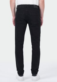 Une personne portant un jean slim noir et des baskets blanches, vue de dos sur un fond clair uni.