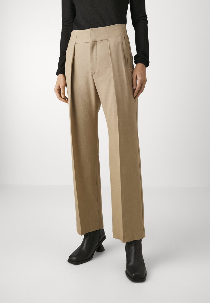 Beige wijde broek met voorplooien, voorzien van een op maat gemaakte tailleband en zijzakken, gestyled met zwarte gehakte laarzen.