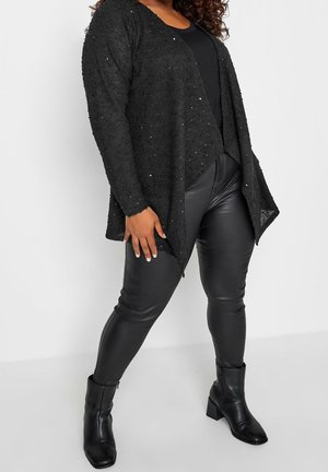 Personne curvy portant un cardigan noir texturé ouvert sur le devant, un haut noir, un pantalon en simili cuir noir et des bottines à talons noires, debout avec les jambes écartées.