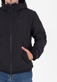 Man in een zwarte hoodie met rits, hand in de zijzak, en blauwe jeans tegen een witte achtergrond.