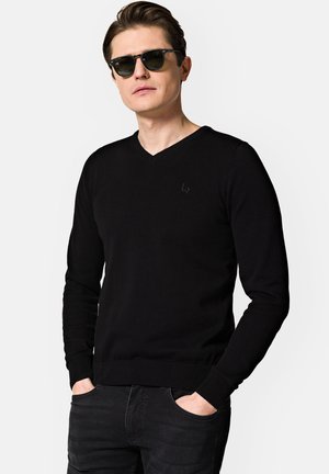 Czarny sweter z dekoltem w serek, wykonany z gładkiego dzianinowego materiału, z długimi rękawami, ribbami na mankietach oraz małym logo na piersi. Noszony z ciemnymi dżinsami.