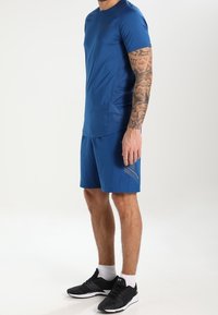 Modrá sportovní košile, odpovídající modré šortky s texturovaným vzorem a černé běžecké boty. Tkanina vypadá lehce a hladce.