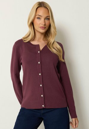 Donna bionda che indossa un cardigan viola con bottoni di perla e pantaloni blu scuro, in piedi davanti a uno sfondo chiaro uniforme.
