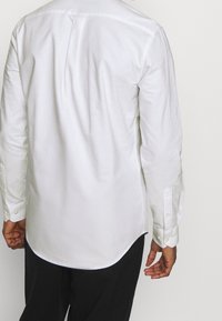 Muž v bílé košili s dlouhým rukávem a černých kalhotách, zobrazen zezadu na jednoduchém světlém pozadí.