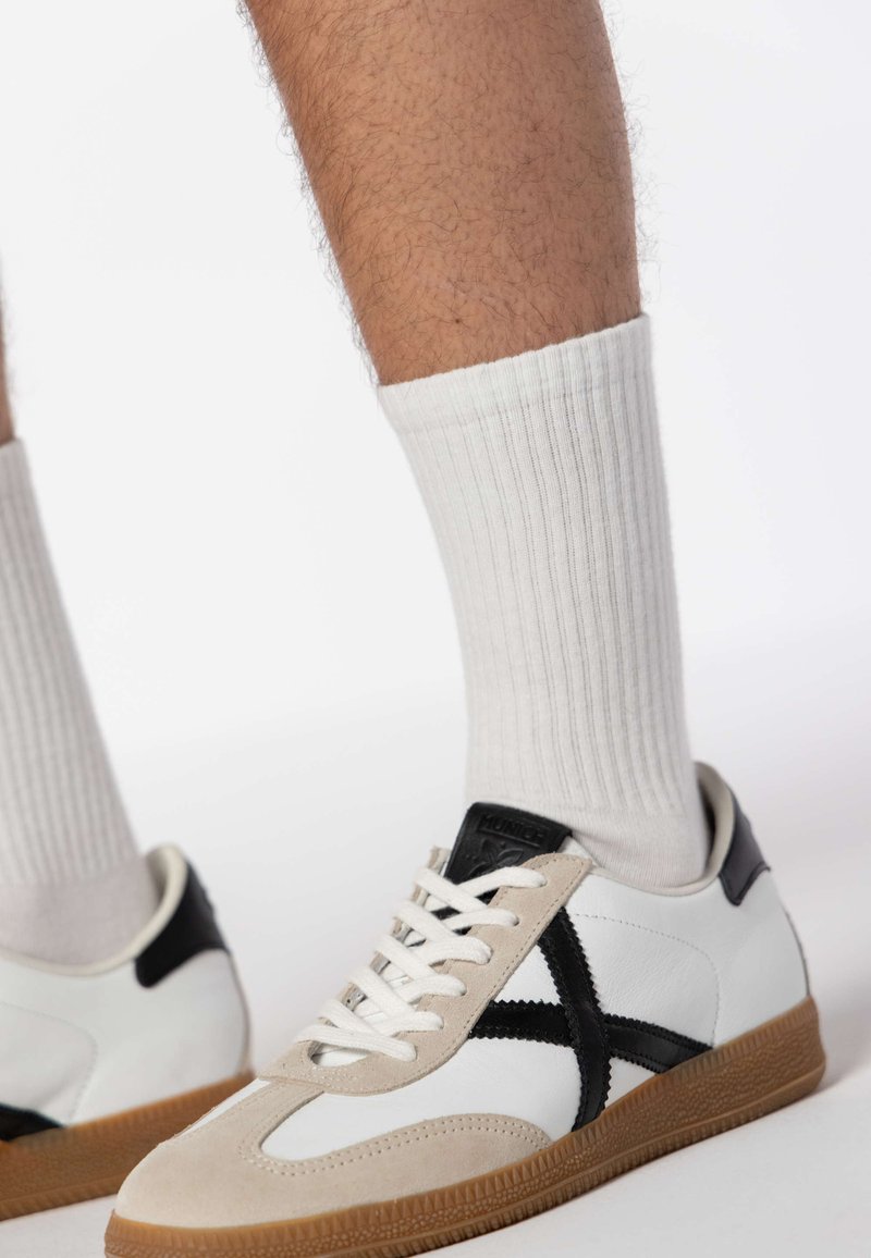 White sneakers with black accents, beige suede details, and a textured rubber sole, paired with white ribbed socks.