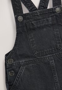 Salopette en denim noir avec bretelles réglables, boutons décoratifs et poche avant. Présente un tissu texturé et des détails de couture robustes.