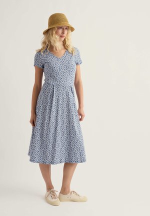 Kobieta w niebiesko-białej sukience w kwiaty do kolan, z beżowym plecionym kapeluszem i beżowymi tenisówkami na sznurowadła, stoi na tle prostym.