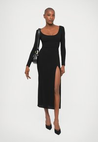 Frau in einem schwarzen langärmeligen Kleid mit einem hohen Schlitz, schwarzen spitzen Absätzen und einer schwarzen Schultertasche, vor einem weißen Hintergrund stehend.