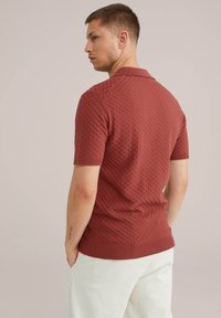 Polo majica s kratkimi rokavi v rdečkasto rjavi barvi, z strukturiranim vzorcem, rebrastim ovratnikom in robom, v kombinaciji z svetlimi hlačami.