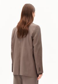 Persoon met lang bruin haar draagt een taupe blazer en bijpassende broek, met de rug naar de camera tegen een effen lichte achtergrond.
