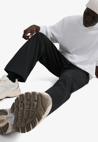 Čierne nohavice v kombinácii s voľnou bielou košeľou s dlhým rukávom a svetlými teniskami. Podrážka tenisiek zobrazuje detailné vzory protišmykovej podrážky.