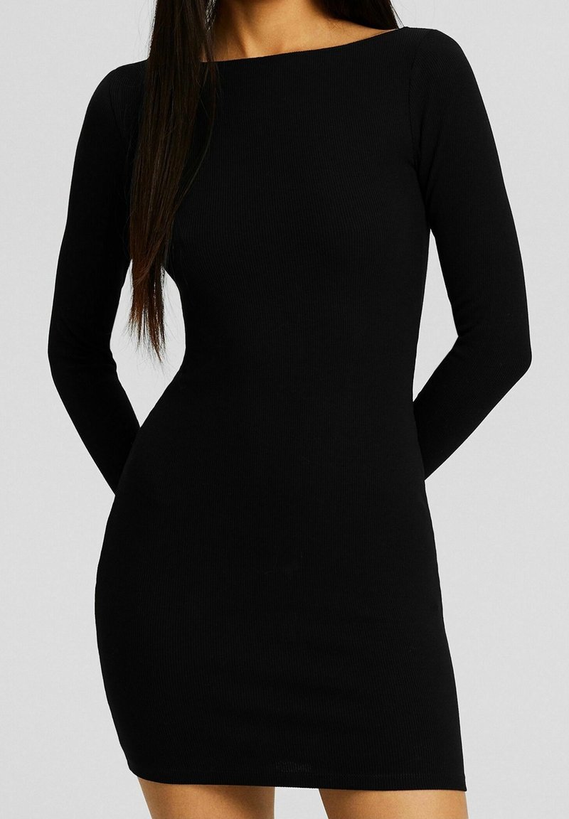 Femme portant une robe mini noire ajustée à manches longues avec un col rond, sur un fond uni.