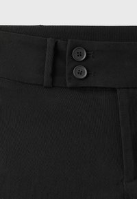 Gros plan de la ceinture d’un pantalon noir montrant deux boutons noirs et une boucle de ceinture sur la texture du tissu.