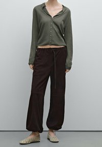 Mørkegrøn knappet cardigan i cropped længde, kombineret med løstsiddende, mørkebrune bukser med snøre. Beige flade sko med sølvdetaljer.