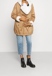 Osoba nosící béžový kabát s kožešinovým límcem přes nosítko pro děti, světle modré zkrácené džíny a černé kotníkové boty, stojící na bílém pozadí.